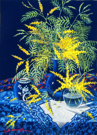 Yellow ball wattle flowers are in a deep blue vase, two other containers are nearby. The whole arrangement stands on a tablecloth with a complicated pattern.