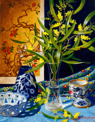 Wattle flowers are in a glass vase. On the wall behind the vase is an oriental scene. A ceramic showing a dragon motif is also present.